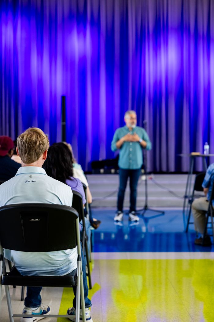 about-01 A speaker addressing a seated audience in a contemporary conference setting with a vibrant backdrop.
